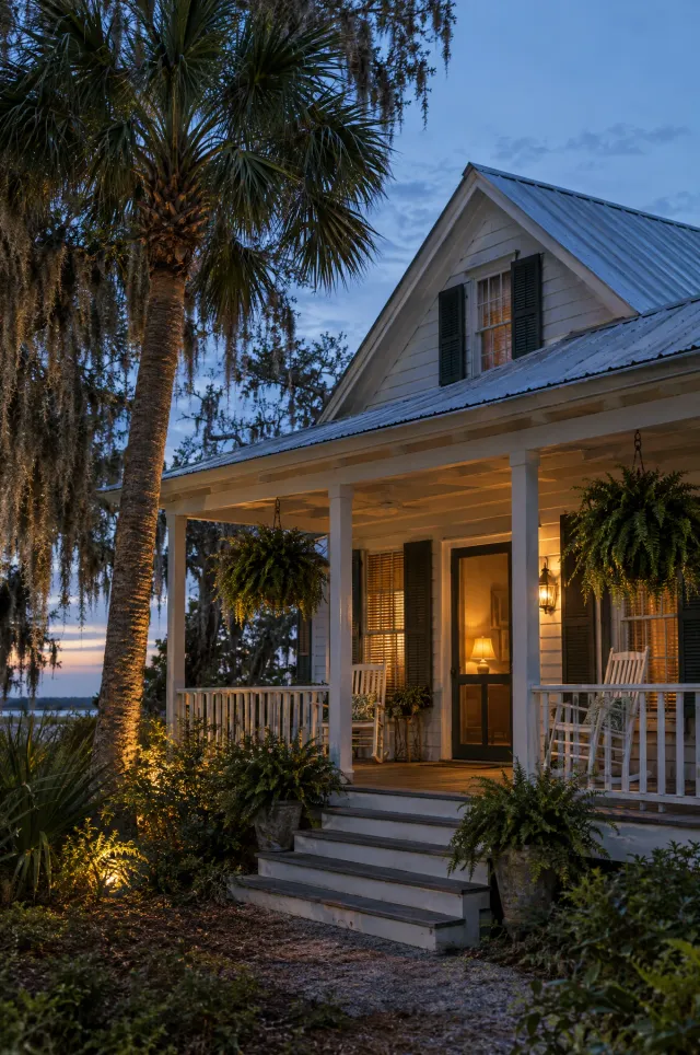 Old Florida cottage porch with rocking chairs, hanging ferns and warm lamplight at dusk beside a palm