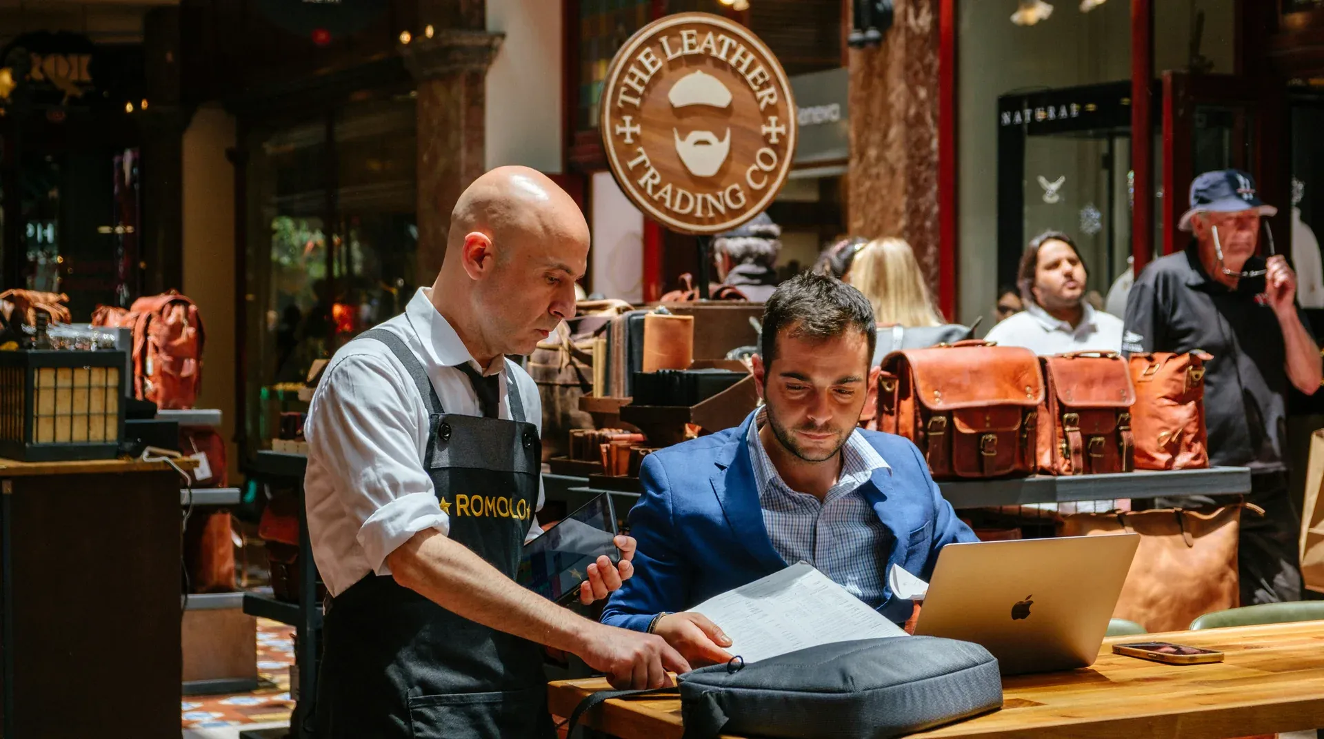 An older shop owner and a younger successor reviewing papers and a laptop in their store