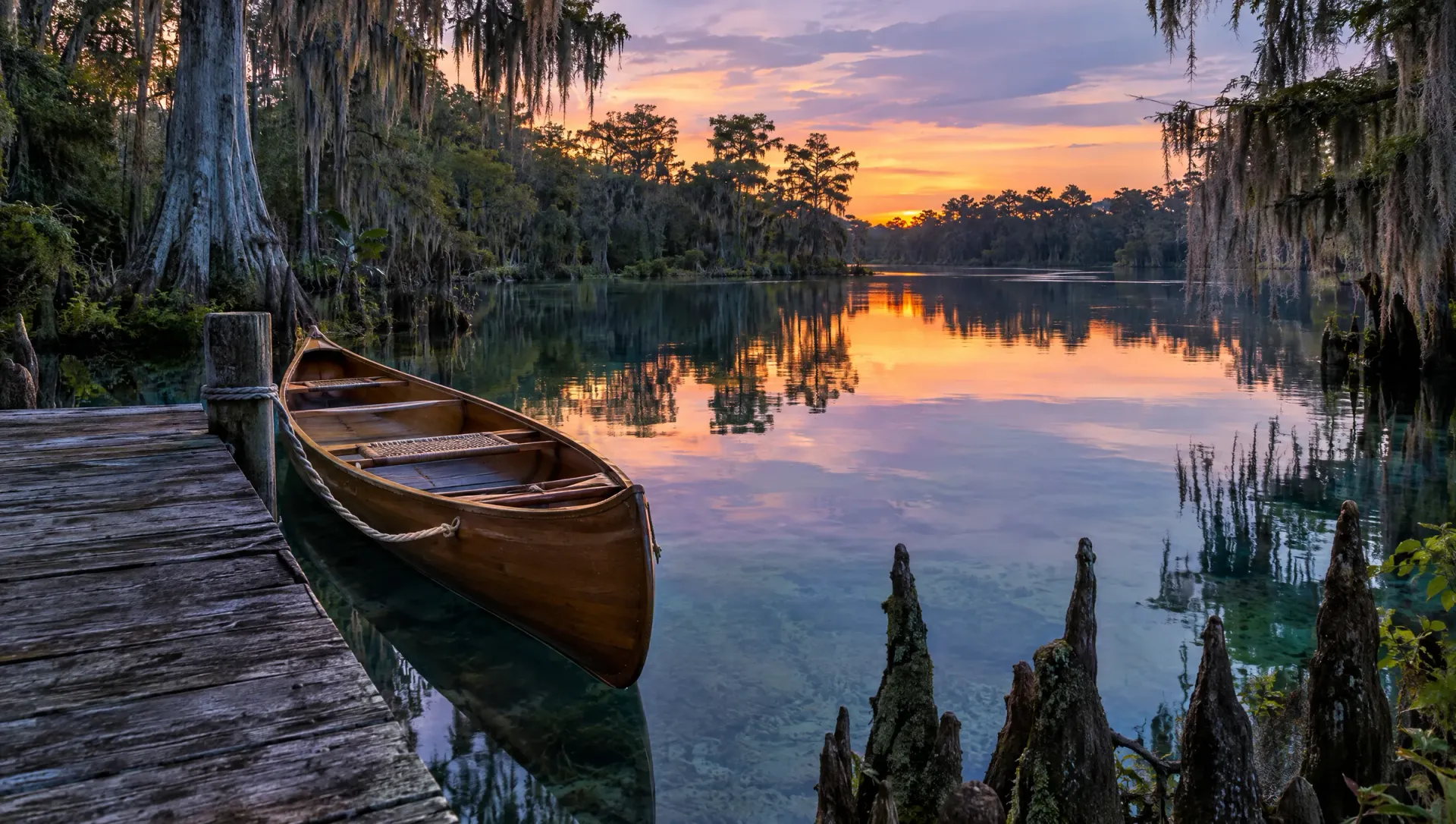 Wooden canoe tied to a weathered dock on a clear cypress-lined Florida spring river at dusk